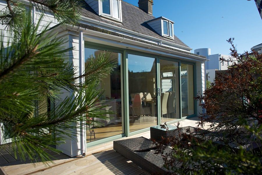 White-clad house with sage green sliding patio doors opening onto a raised timber deck, surrounded by garden plants and outdoor seating.